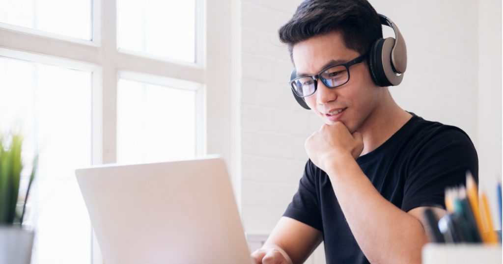 Closeup of a young man sitting at a desk wearing headphones, looking at his laptop.