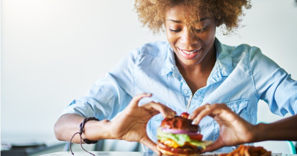 Closeup of a woman sitting at a table holding a burger in her hands with a plate of fries underneath.